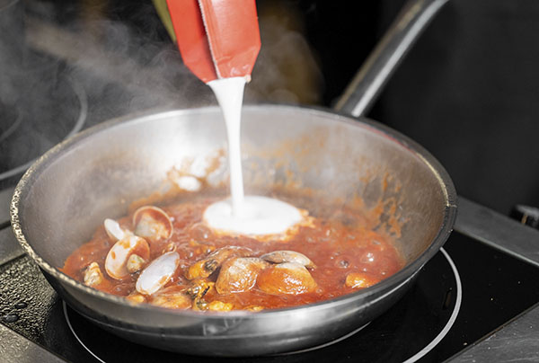 Kokki lisää ruokakermaa pannulle simpukoita sisältävään ruokaan.